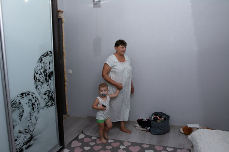 In a softly lit room, a grandmother in a simple white dress holds hands with her grandson, dressed in a tank top and shorts. The setting is a lived-in interior, offering a glimpse into family life.の写真素材