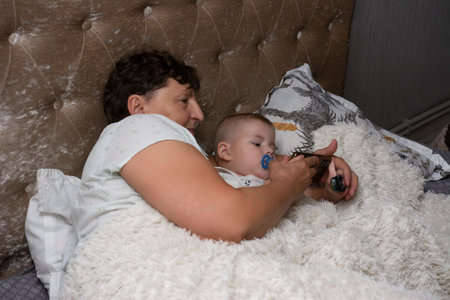 A grandmother and baby lie together in bed, watching a phone screen. The baby has a pacifier. The bed has a soft blanket. This is a bonding moment with technology at the forefront.の写真素材