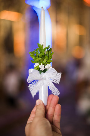 A close-up shot of a hand holding a white candle adorned with greenery and a lace ribbon. The background is blurred, suggesting a ceremonial or celebratory setting.の写真素材