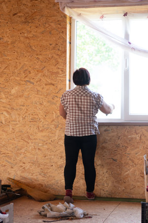 A woman stands with her back to the viewer as she installs a window. She is wearing a plaid shirt and black pants. The wood paneled room is sparse with brooms and rags on the floor.の写真素材