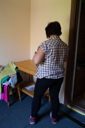 Back view of a woman in a plaid shirt standing in the corner of a room. A wooden table with a pile of laundry sits nearby. She is wearing black pants and purple shoes. The setting is domestic.の写真素材
