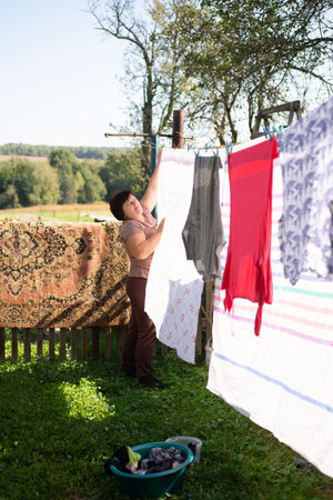 This image shows a woman hanging up laundry on a clothesline in her backyard on a clear, sunny day. A colorful rug hangs on the fence behind her. A wash basin sits on the grass.の写真素材