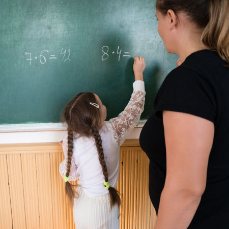 A young student, back to camera, solves a math problem on a green chalkboard, aided by a teacher observing to the right. The classroom setting promotes education and learning.の写真素材