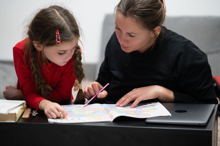 A mother and daughter are sitting together at a black table, working on a school project. They are...
