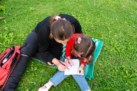 A woman and a child sitting together on a blanket on the grass, drawing in a notebook. Both are focused on their artwork, enjoying a peaceful and creative moment outdoors.の写真素材