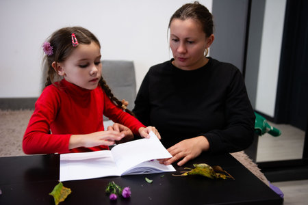 In a brightly lit interior, a young girl in a red shirt is reading an album of pressed plants and...