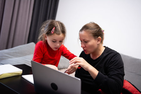 In a domestic setting, a mother supports her child with homework, employing a laptop for educational activities. A notebook is present on the black table near the couch.の写真素材