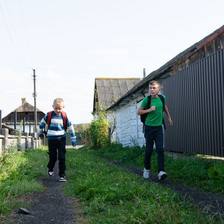 Two young boys are walking on a dirt path. They are wearing backpacks, suggesting they are students. The houses are aged, adding to the rustic feel of the setting. The sun shines.の写真素材