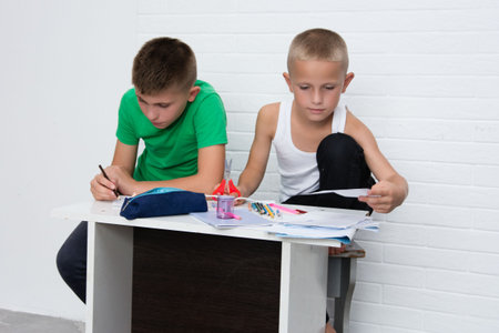 Captured indoors, the image features two young boys engaged in drawing. One boy, in a green t-shirt, is actively sketching. His companion, dressed in a white tank, examines a paper. Creative moment.の写真素材