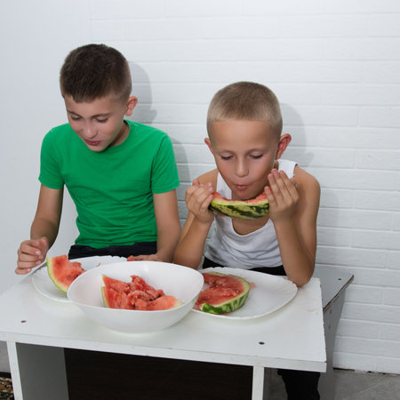 This picture shows two young boys sitting at a table, heartily enjoying slices of watermelon. The joyful scene against a white brick wall depicts friendship.の写真素材
