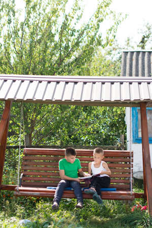 Two young boys are sitting together on a wooden swing, captivated by a book they're reading. They're outdoors on a sunny day, surrounded by lush greenery, creating a peaceful atmosphere.の写真素材