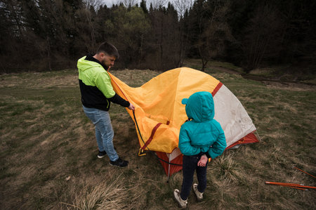 Setting up a bright tent in a lush green area, an adult and a child share a delightful moment, embracing nature and preparing for an adventurous night under the stars.の写真素材