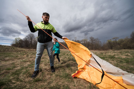 On a cloudy afternoon, a man skillfully assembles a colorful tent while a child in a bright jacket gleefully watches nearby, ready for an exciting outdoor experience.の写真素材