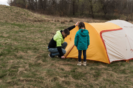 A child in a turquoise jacket watches as an adult carefully secures the yellow and orange tent on...