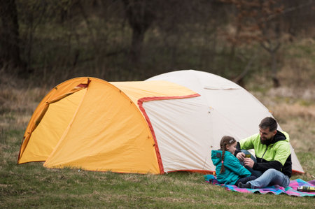 A father and daughter share a joyful moment outside their yellow tent, surrounded by nature's beauty, creating lasting memories on a serene day.の写真素材