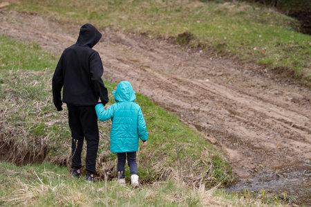 Kids enjoy a serene moment by a small stream, holding hands and discovering the beauty of nature on a cool spring day, surrounded by fresh grass and trees.の写真素材