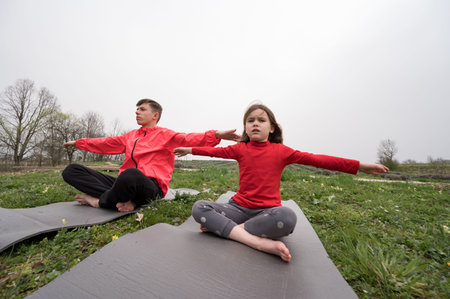 Two children practice yoga on mats in an open field, surrounded by lush grass and trees. They demonstrate concentration and balance amidst the gentle sounds of nature.の写真素材
