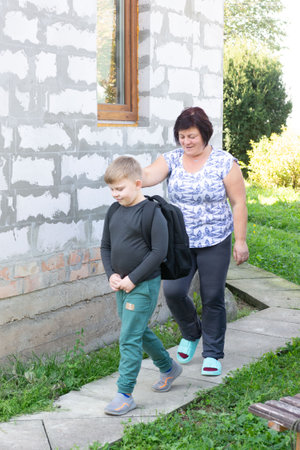 A grandmother walks with her grandson to school, placing a hand on his shoulder. They are on a concrete path near a building under construction with a lush green backdrop.の写真素材