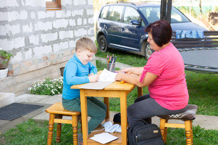 A boy is sitting at a table with his grandmother, drawing a picture together outside in the yard next to a house. A trampoline is in the background.の写真素材