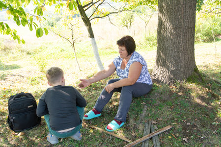 A grandmother sits next to a large tree in a grassy field, sharing a moment with her grandson who is crouched nearby. It is a sunny day outdoors, in a tranquil countryside setting.の写真素材