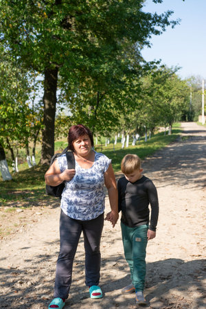A woman and a young boy are walking hand-in-hand along a dirt path lined with trees. The woman is wearing a printed top and carrying a backpack. The boy looks downcast.の写真素材