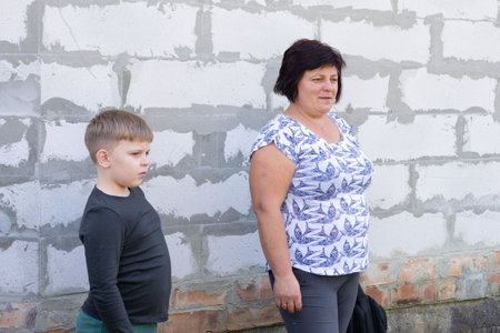 This outdoor shot features a woman and a young boy standing side-by-side against a cinderblock wall. The mother wears a bird print shirt and gray pants. The wall features both cinder blocks and brick.の写真素材