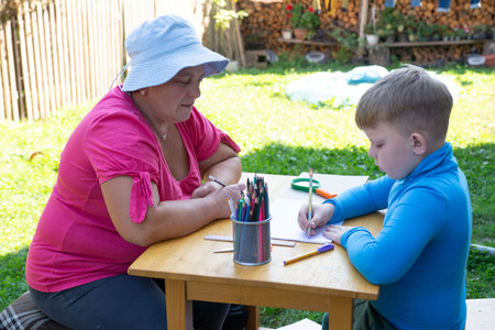 This image captures a child drawing at an outdoor table with an adult assisting or observing. Surrounded by a vibrant garden, it's a snapshot of learning and creative expression in a natural setting.の写真素材