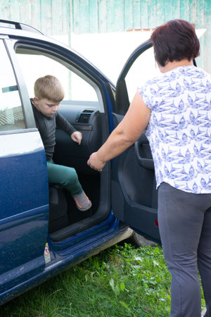 A young boy carefully enters a blue car, with an adult holding the door open for him. The setting appears to be outdoors, with a rustic wooden fence in the background and grass nearby.の写真素材