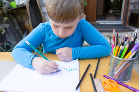 A young boy in a blue shirt sits outdoors, drawing with colored pencils. The child is focused on landscape art. The wooden table has charcoal, pencils, ruler, and scissors.の写真素材