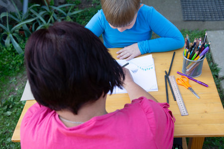 Captured from an overhead perspective, the image showcases a woman instructing a child on drawing. They are seated outdoors at a wooden table filled with art supplies, promoting creativity.の写真素材