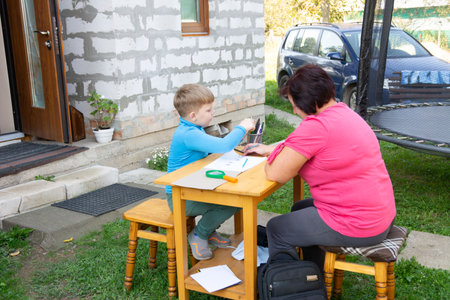 A young boy and an adult woman are seated at a small wooden table outside, deeply engaged in a learning activity. The pair are working on a table on the lawn near the house.の写真素材