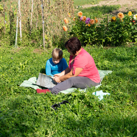 In a sun-drenched garden, a woman and a child are absorbed in a book while sitting on a blanket. The scene is peaceful and idyllic, surrounded by plants and flowers.の写真素材