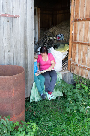 A woman and a child are sitting together outside a weathered barn, engrossed in a book. The scene exudes rural simplicity, framed by lush greenery and rustic architecture.の写真素材