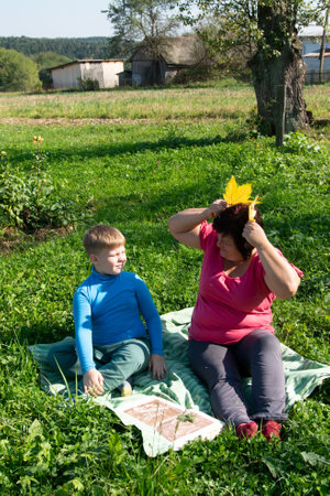 A boy and a woman are picnicking outside, the woman is creating a crown out of yellow leaves. They sit on a blanket in a meadow, surrounded by green grass, with a rustic building in the background.の写真素材