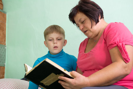 A woman reads a book to a boy. They are sitting on a bed against a green wall. The boy is looking at the book with attention as the woman reads aloud to him in the room.の写真素材