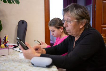 In a warmly lit room, a grandmother and her granddaughter are seated at a table, absorbed in their smartphones. They share space, yet their attention is captivated by digital interaction.の写真素材
