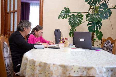 An older woman helps a girl with her schoolwork. They are seated at a table in a home setting. There's a large plant, a laptop, and school supplies on the table. An open door is in the background.の写真素材