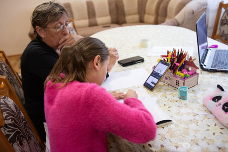 A grandmother attentively supervises her granddaughter as she focuses on completing her homework. The student uses her phone and laptop as learning aids at the round kitchen table.の写真素材