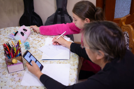 An image depicts a girl and an adult working on a drawing at a table. The scene includes drawing supplies, a mobile phone in use, and instruments behind, suggesting a creative environment.の写真素材