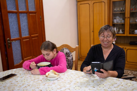 A young girl eats soup at a table while an older woman watches something on a handheld device. This cozy setting reflects warm family moments, togetherness, and simple home life.の写真素材
