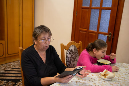 Indoor scene shows an older woman using a smartphone and a young girl eating soup. The room is warmly lit, with a table, patterned tablecloth, and wooden furniture.の写真素材