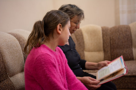 In an indoor setting, a grandmother and granddaughter are seated closely together on a sofa, deeply engrossed in reading a book. The grandmother is holding the book open.の写真素材