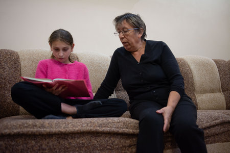 A young girl reads from a book with a red cover while sitting cross-legged on a brown sofa. An older woman with glasses sits next to her. The scene suggests intergenerational bonding and storytelling.の写真素材