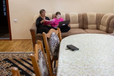A grandmother and granddaughter are sitting together on a couch. The grandmother is reading a book, and the granddaughter is working on a laptop indoors in a cozy, neutral toned room.の写真素材