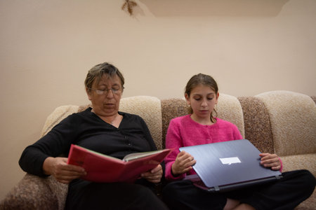 An older woman with glasses sits on a couch reading a book. A young girl sits next to her, using a laptop computer. Both are focused and engaged in their respective activities.の写真素材