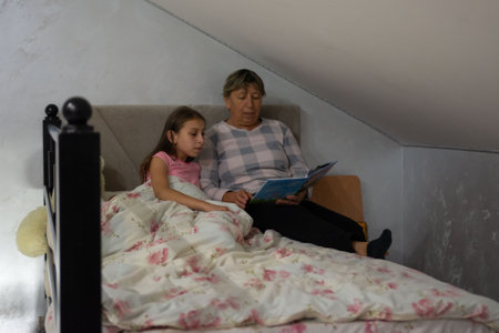 A heartwarming scene captures a grandmother reading to her granddaughter in bed. The room is cozy, featuring a comfortable bed covered in a floral print. The setting promotes familial bonding.の写真素材