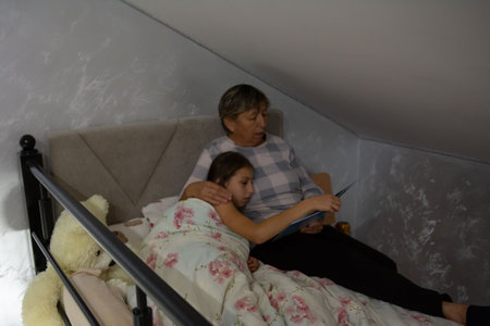A grandmother shares a story with her granddaughter in a cozy attic bedroom. The girl is wrapped in floral bedding, while a teddy bear rests nearby, creating a warm and familial scene.の写真素材