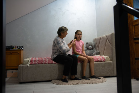 This image shows a grandmother carefully combing her granddaughter's hair. They are seated side-by-side on a bed with a teddy bear nearby in a cozy, casual home setting.の写真素材