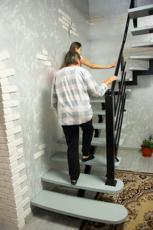 A woman and a girl are going up a set of floating stairs with gray steps and a black railing. The surrounding space features textured gray and white walls and a patterned carpet.の写真素材