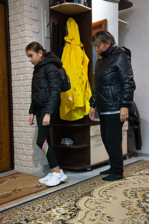 Two people, an adult and child, stand in the entryway of a home. The girl wears black leggings with colored stripes and a black jacket. The adult wears black pants and a black puffy coat.の写真素材